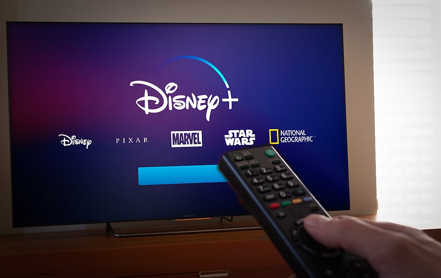 Person pointing remote control at Disney+ on big screen TV with Disney, Pixar, Marvel, Star Wars and NatGeo logos displayed