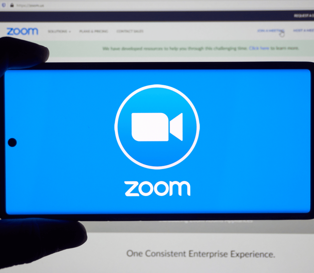 Person holds smartphone with Zoom logo in front of a desktop monitor with the Zoom application open