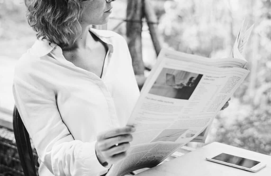 Black and white photo depicting a business woman reading the newspaper with a smartphone on the table in front of her