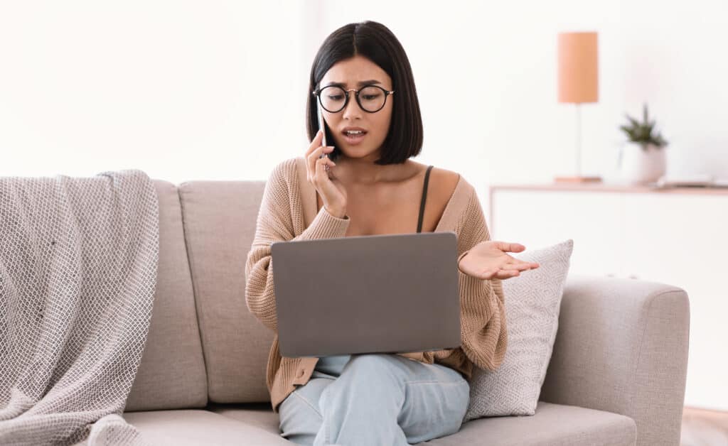 Frustrated serious woman in glasses talking on mobile phone, sitting on couch and using laptop, having problem with internet connection, calling customer support hotline