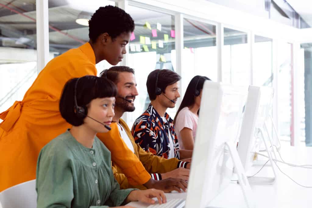 Side view of diverse customer service executive trainer assisting her team at desk in office