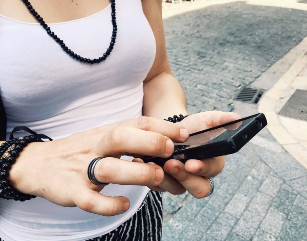 Young woman wearing white top and black jewelry types on smartphone, signifying customers who post frustration to social media.