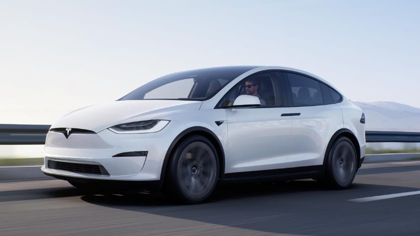 White Tesla Model X being driven by a man on a highway with mountains in the background