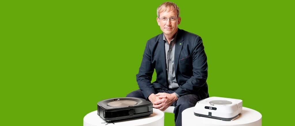 iRobot CEO Colin Angle sits behind two iRobot products in front of green background.