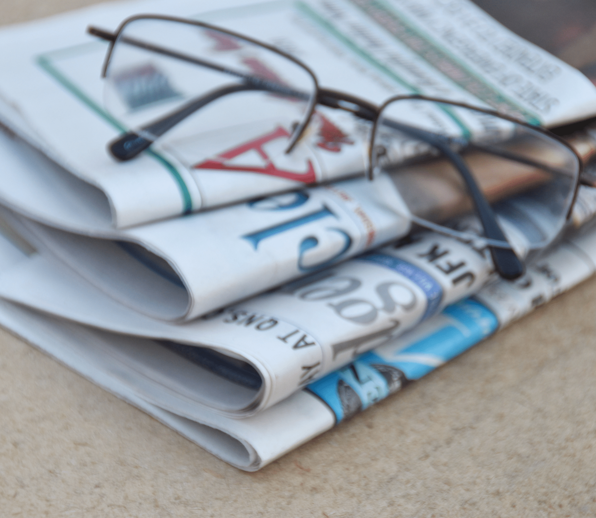 A pair of wire-rimmed glasses sits on top of four folded newspapers on a table.