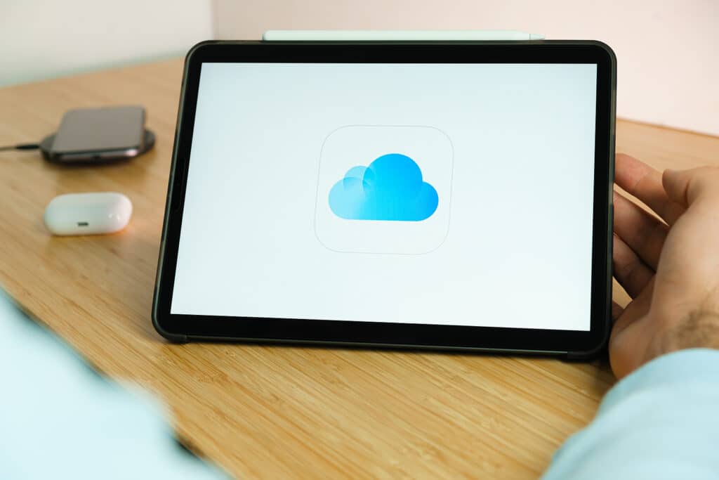 ICloud logo on the screen of iPad tablet with smart phone iPhone charging on the wireless charger and airpods in the case on the background