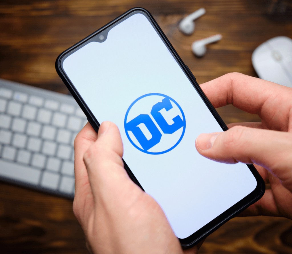 DC logo shown on a smartphone, held above a white keyboard and earbuds