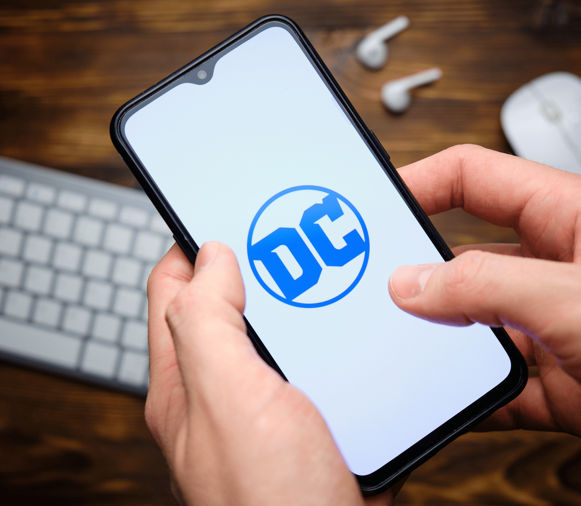 DC logo shown on a smartphone, held above a white keyboard and earbuds