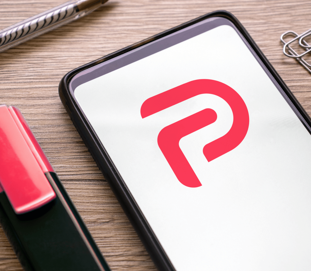 Red Parler logo on a white background on a smartphone, sitting on a desk between a red marker and paper clips