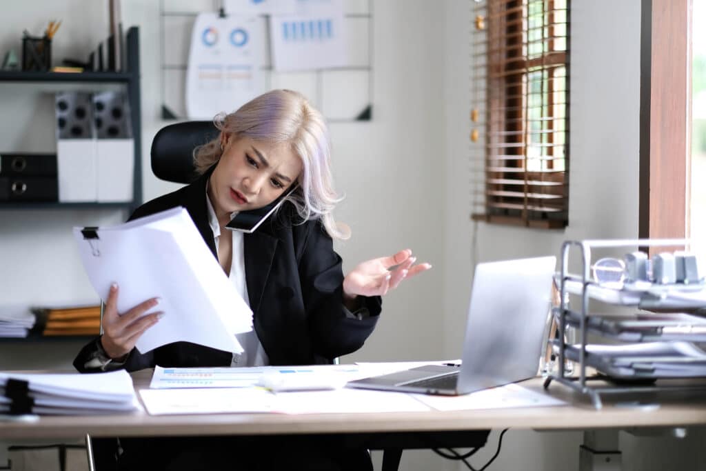 Asian woman cover her face with hand and feel upset while talk on mobile phone with customer in front of laptop computer on desk at office, illustrating the frustration of Vonage customers who couldn't cancel their service due to dark patterns.