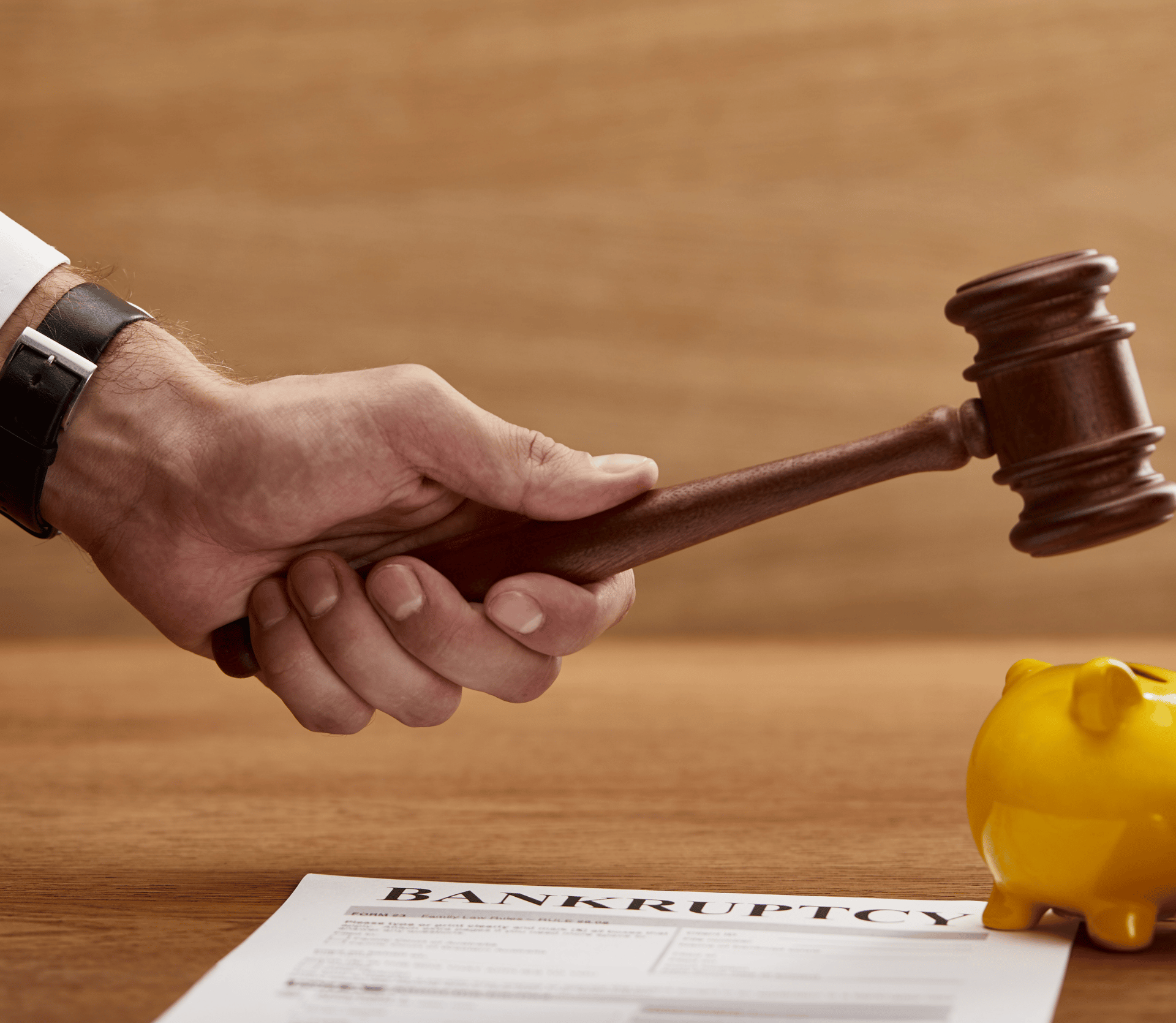 Man holding gavel over a yellow piggy bank, with a document with the word BANKRUPTCY typed at the top.