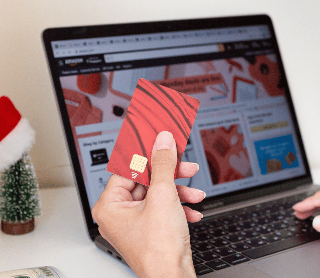 Woman shopping at Amazon on her laptop, holding a red credit card with a Christmas tree and Santa hat in the background.