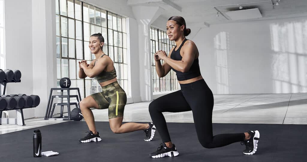 Two women doing strengthening exercises in a brightly lit studio with white walls, a black and white floor with weights on the lef hand side.