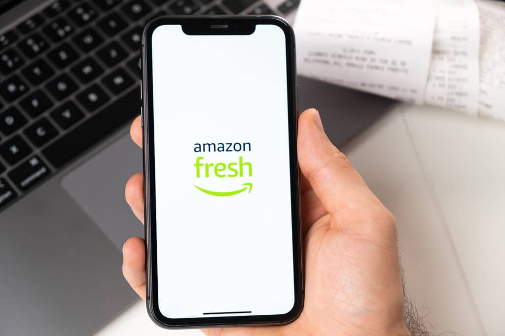 Amazon fresh application of supermarket and grocery store in a smartphone. A man is holding a smartphone with app for shopping and laptop on the background.