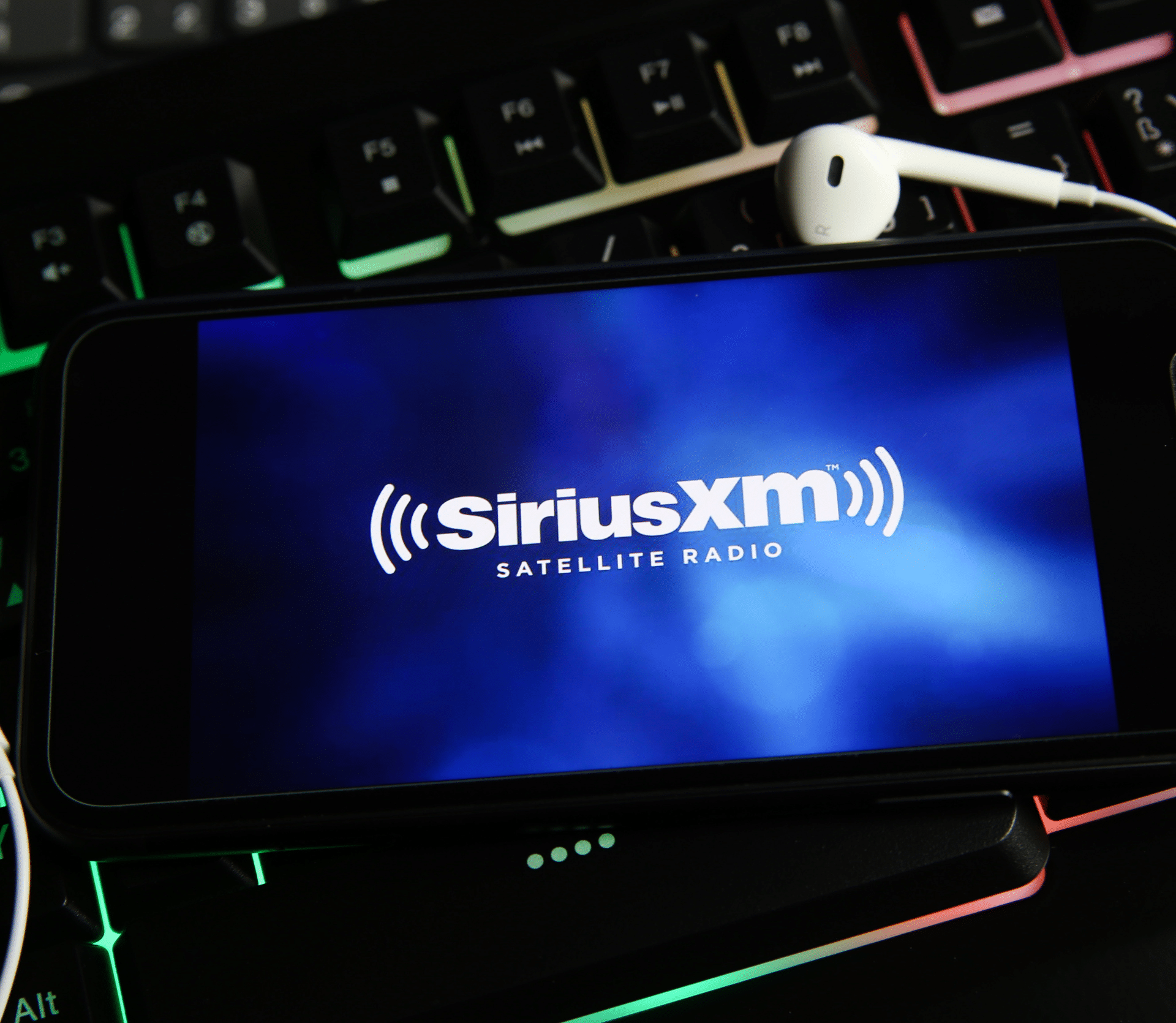 SiriusXM logo displayed on smartphone with backlit keyboard and headphones as background images