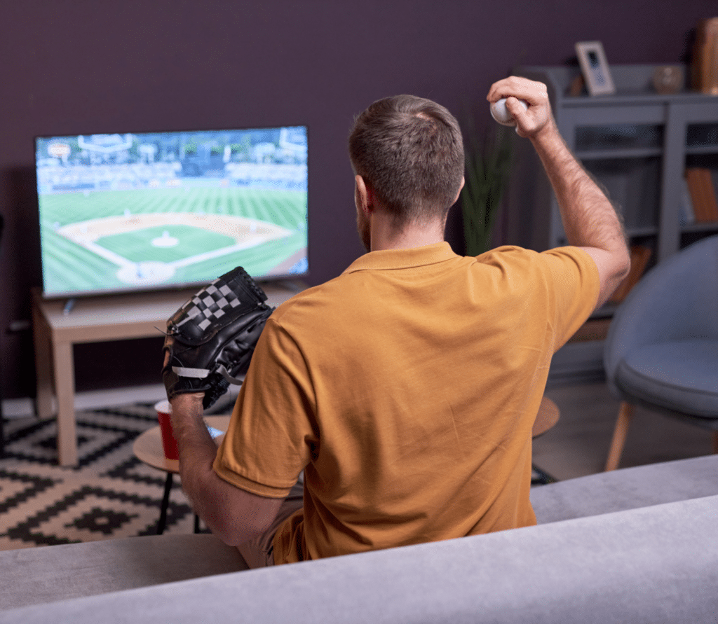 Man holding baseball and glove watches baseball game on TV.