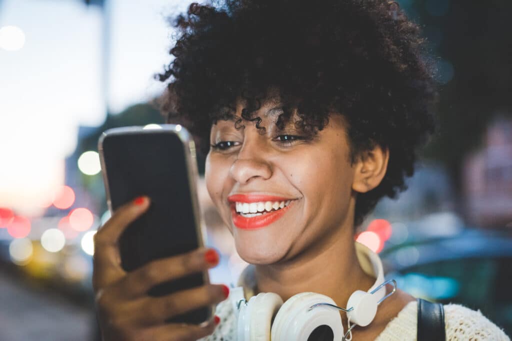 Portrait of young handsome afro woman holding a smartphone, looking the screen smiling - social network, communication, technology concept - face illuminated by screen light