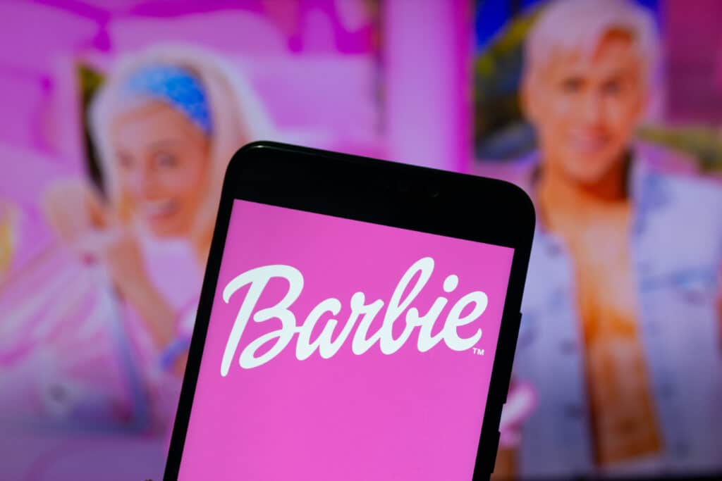 The Barbie logo with a pink background on the screen of a smartphone with the actors of the film in the background.