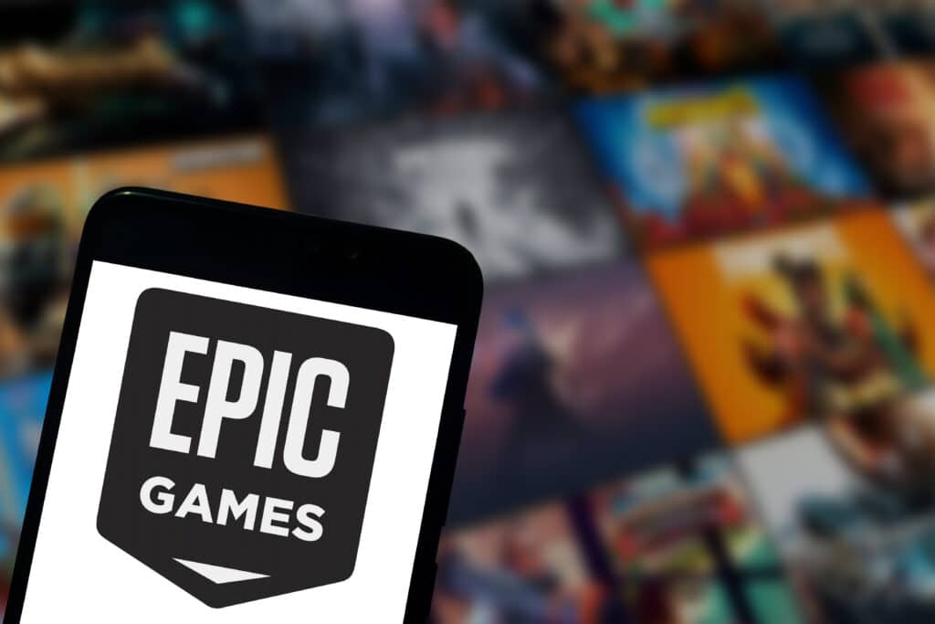 The Epic Games logo is displayed on a smartphone screen with a blurred background of a TV screen in the background with various games from the company.